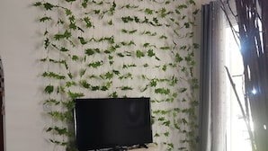 Een woonkamer met een open haard, een televisie, een gestreept fauteuil en een decoratieve wand met hangende bladeren.