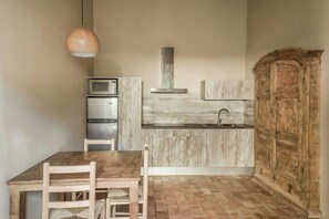 A compact kitchen with a wooden table, chairs, a microwave, a refrigerator, a sink, and a rustic cabinet. - Flat in a former rectory near Begur (Regencós, Girona)