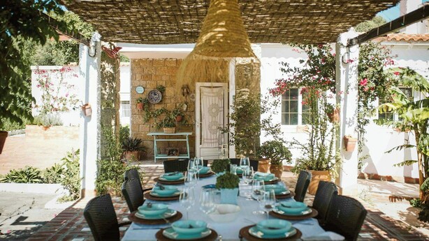 An outdoor dining area with a table set for six, surrounded by chairs, under a pergola with a hanging light fixture. - Centuries-old olive finca with pool in Cádiz (Barbate)