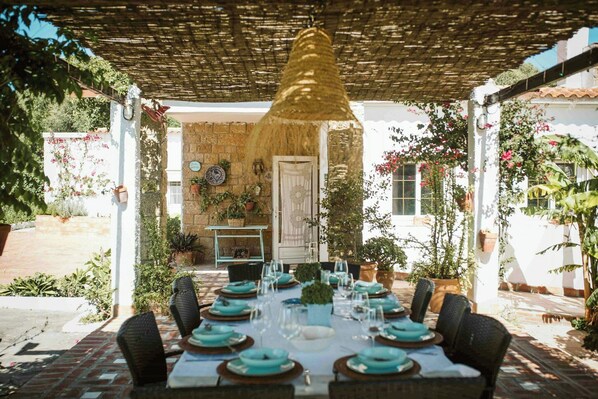 Ein Essbereich im Freien mit einem Tisch für sechs Personen, umgeben von Stühlen, unter einer Pergola mit einer hängenden Leuchte.