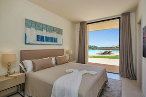 A bedroom with a large bed, a painting on the wall, a nightstand with a lamp, and a view of a pool and lounge chairs through an open door.