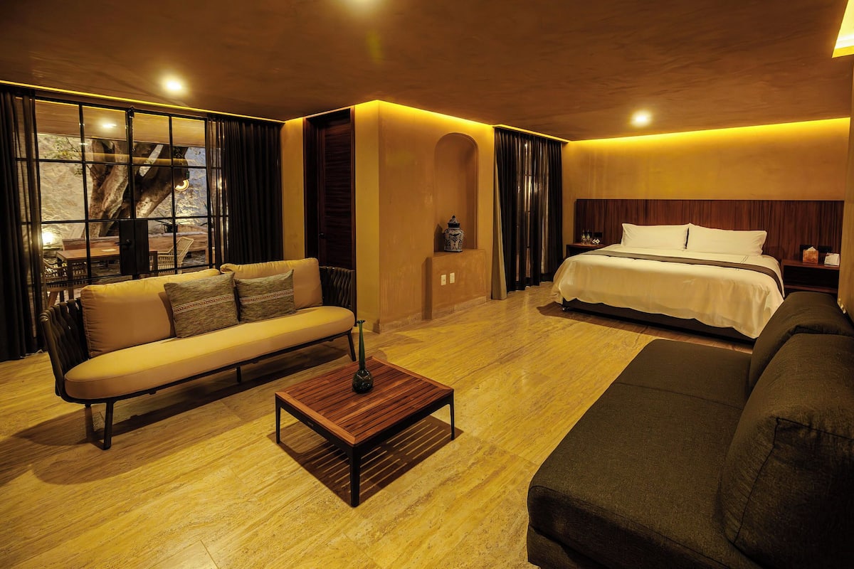 Quarto de hotel moderno com sofá, cama, mesinha de madeira e vista para uma árvore através de grandes janelas.