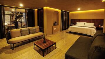 A modern hotel room with a sofa, a bed, a wooden coffee table, and a view of a tree through large windows.