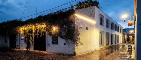 A cobblestone street with a building featuring a white facade, a vine-covered wall, and a lit sign.