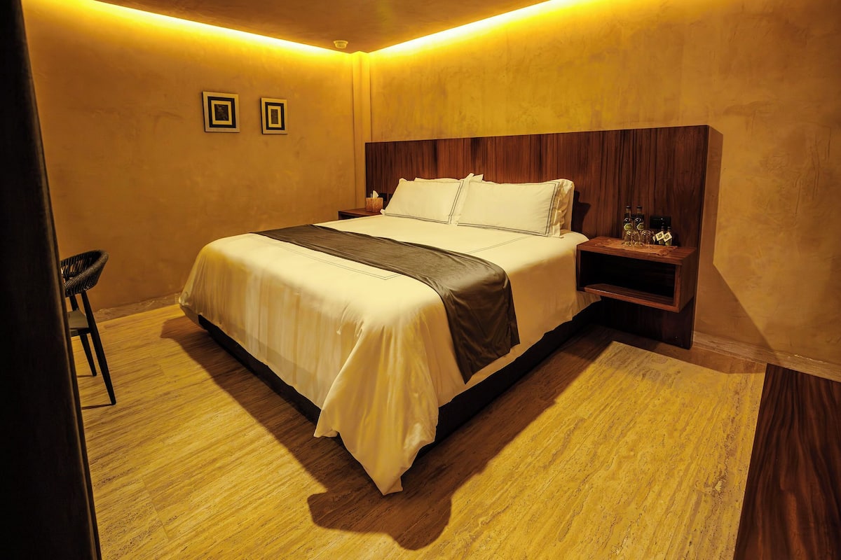 Uma cama bem arrumada com cabeceira, uma mesa de cabeceira com abajur e uma cadeira.