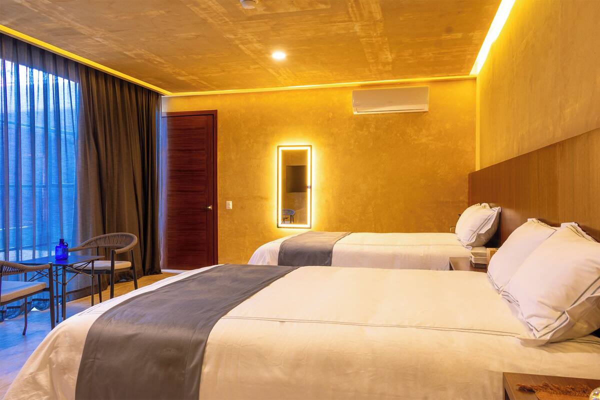 Quarto de hotel com duas camas, uma escrivaninha com cadeira, uma janela grande com cortinas e um espelho com iluminação.