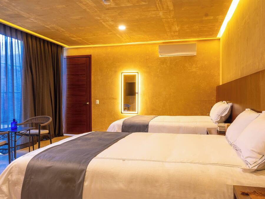 Habitación de hotel con dos camas, un escritorio con silla, un ventanal con cortinas y un espejo con iluminación.