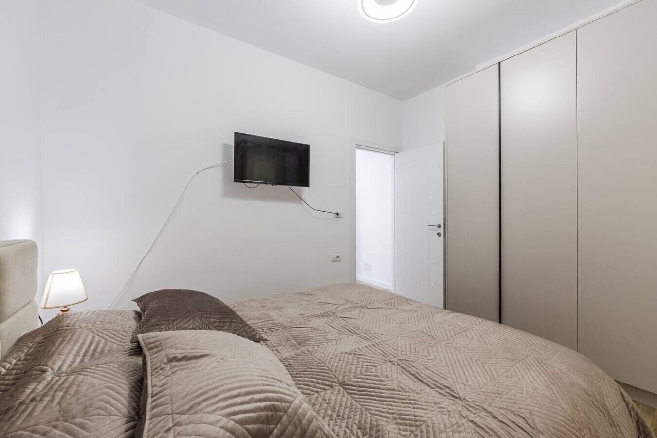 Un dormitorio con cama, televisor montado en la pared, armario y puerta que da a otra habitación.