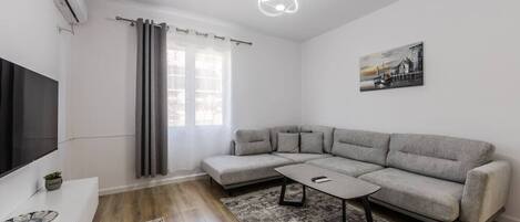 Una sala de estar moderna con un sofá seccional gris, un televisor de pantalla plana, una mesita y un ventanal con cortinas.
