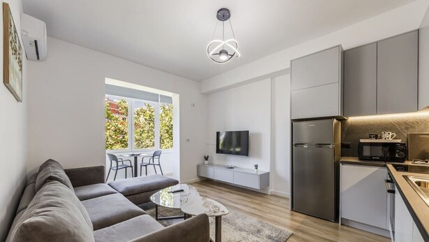 A modern apartment with a grey sofa, a small coffee table, a TV, a kitchen with a fridge and microwave, and a balcony with a view of greenery. - Central Tirana 1BR Apartment Near Main Square by PikHost (Tiranë)