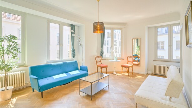 Een woonkamer met een blauwe bank, een witte bank, een glazen salontafel, een houten stoel en een hanglamp.