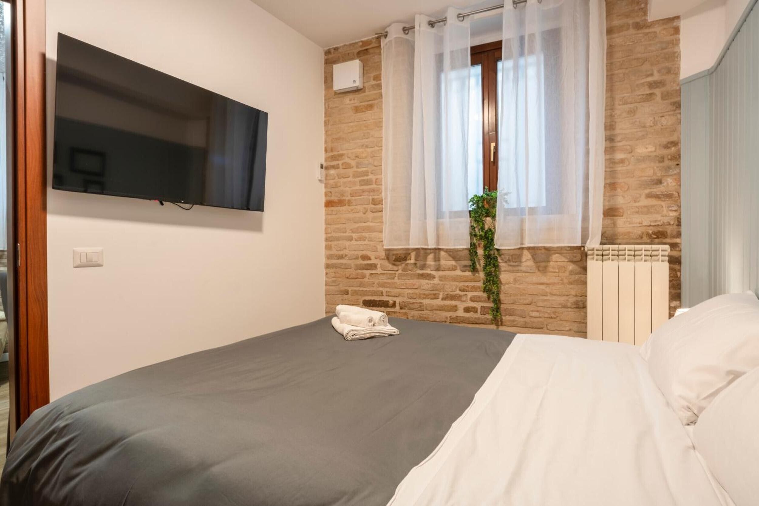 Une chambre à coucher comprenant un lit, un téléviseur à écran plat, un mur en briques, une fenêtre avec des rideaux légers et un radiateur.