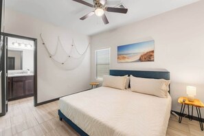 Ein Schlafzimmer mit Bett, Deckenventilator, Nachttisch, Lampe, einem Gemälde sowie einem Badezimmer mit Spiegel und Waschbecken.