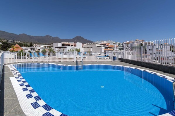 A rooftop swimming pool with clear blue water, surrounded by a white fence, and a view of buildings and mountains in the background. - Costa Caleta 1 - Two Bed (La Caleta)
