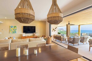 Ein modernes Wohnzimmer mit einer großen Couch, einem Esstisch mit Kerzen, Korbtreleuchten und Blick auf das Meer.