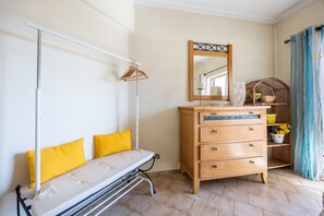 A bedroom with a bed, a wooden dresser, a mirror, a hanging rack, and a window with a curtain. - Apartment 'Sunny Quarteira Escape - Algarve' with Shared Garden, Balcony and Wi-Fi (Quarteira)