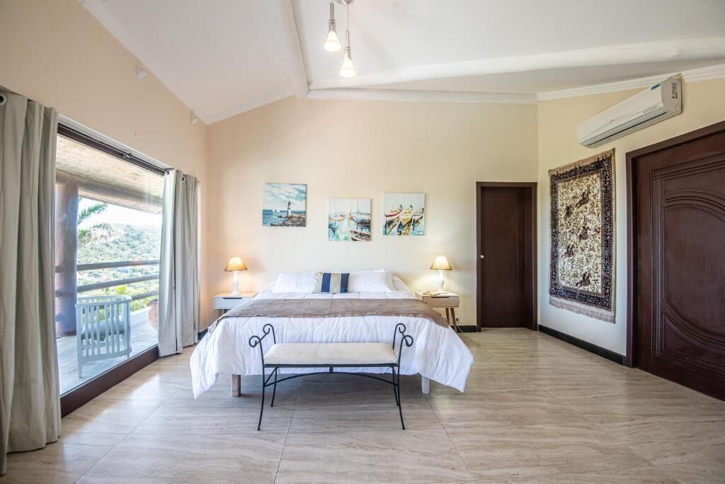 A hotel room with a bed, bedside lamps, a bench, a large window with curtains, and artwork on the walls.
