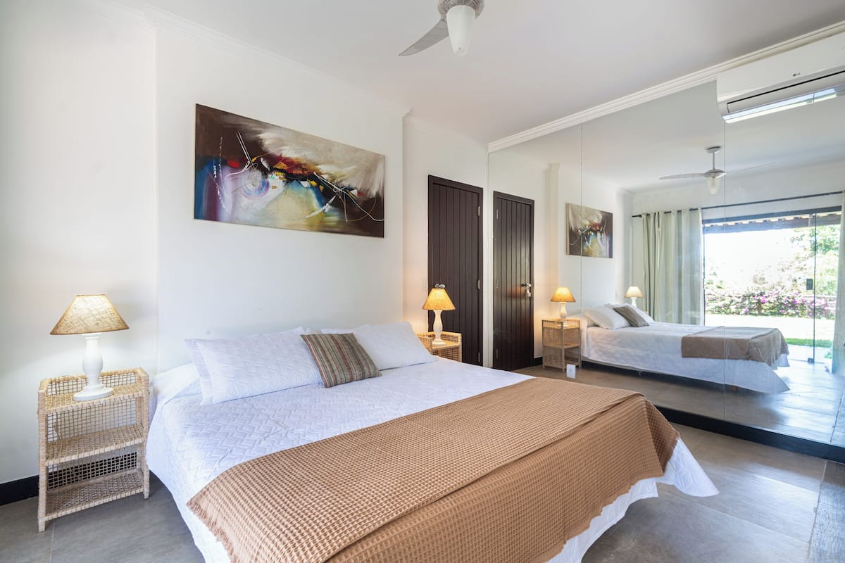 A hotel room with a large bed, two bedside tables with lamps, a ceiling fan, and a large window with a view.
