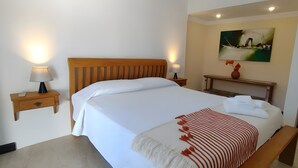 Habitación de hotel con una cama, mesitas de noche, un banco, un cuadro y un jarrón con flores.