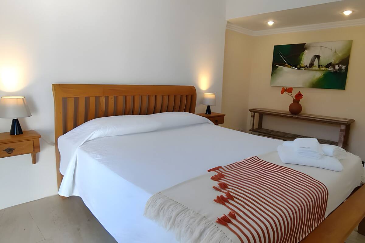 A hotel room with a bed, bedside tables, a bench, a painting, and a vase with flowers.