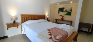 Ein Hotelzimmer mit Bett, Nachttischen, einer Bank, einem Gemälde und einer Vase mit Blumen.
