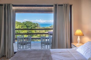 Una habitación de hotel con cama, una mesita de noche con lámpara, y un balcón con sillas y una mesa.
