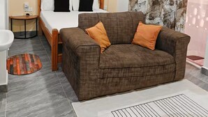 Ein Schlafzimmer mit einem Bett, einem braunen Sofa, einem kleinen Nachttisch, einem runden Teppich und einem Badezimmer mit Waschbecken und Spiegel.