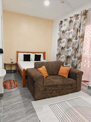A bedroom with a bed, a brown sofa, a small bedside table, a round rug, and a bathroom with a sink and mirror. - Stylish Mtwapa Studio with Rooftop Pool View (Mtwapa)