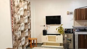 Ein modernes Wohnzimmer mit einer Couch, einem Fernseher, einem kleinen Tisch und einer Küchenzeile mit Schränken und Geräten.