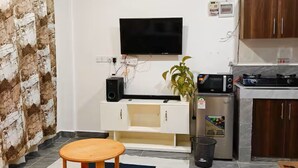 Ein modernes Wohnzimmer mit Flachbildfernseher, Couch, Holztisch und einer Küchenzeile mit Mikrowelle und kleinem Kühlschrank.
