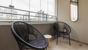 Ein Balkon mit zwei schwarzen Korbstühlen, einem kleinen runden Tisch, einem großen Fenster mit Blick auf Gebäude und einem wandmontierten Spiegel.