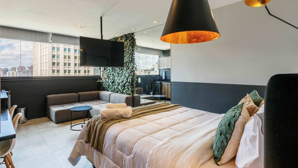 A modern hotel room with a large bed, a sofa, a small table, a TV, and a city view. - Studio Completo a 5min do Metro V Olimpia | VG1703 (Vila Olímpia)