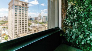 A window with a cityscape view, a green leafy wall, and a window sill. - Studio Completo a 5min do Metro V Olimpia | VG1703 (Vila Olímpia)