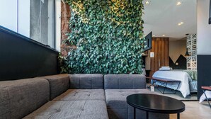 A modern hotel room with a large sectional sofa, a small round table, a bed with a white comforter, and a green leafy wall. - Studio Completo a 5min do Metro V Olimpia | VG1703 (Vila Olímpia)