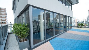 Un edificio moderno con grandes ventanales, un espacio exterior empedrado con baldosas azules y beige, y plantas en macetas.