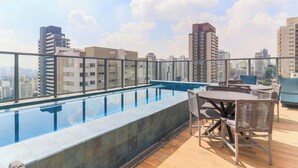 Una piscina en la azotea con vistas a edificios de la ciudad, mobiliario exterior y cielo despejado.