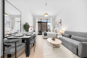 A modern living room with a dining area, grey sofa, and a large window with a city view. - Free housekeeping visits during your stay - StayShort - Beauriful Modern Studio that Sleeps 4 at Riviera Beachfront (Meydan)