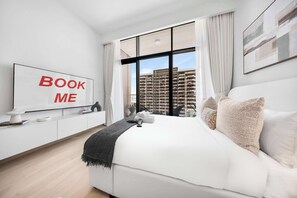 A modern bedroom with a large bed, a mounted TV displaying 'BOOK ME,' a view of high-rise buildings, and a minimalist design. - Free housekeeping visits during your stay - StayShort - Beauriful Modern Studio that Sleeps 4 at Riviera Beachfront (Meydan)