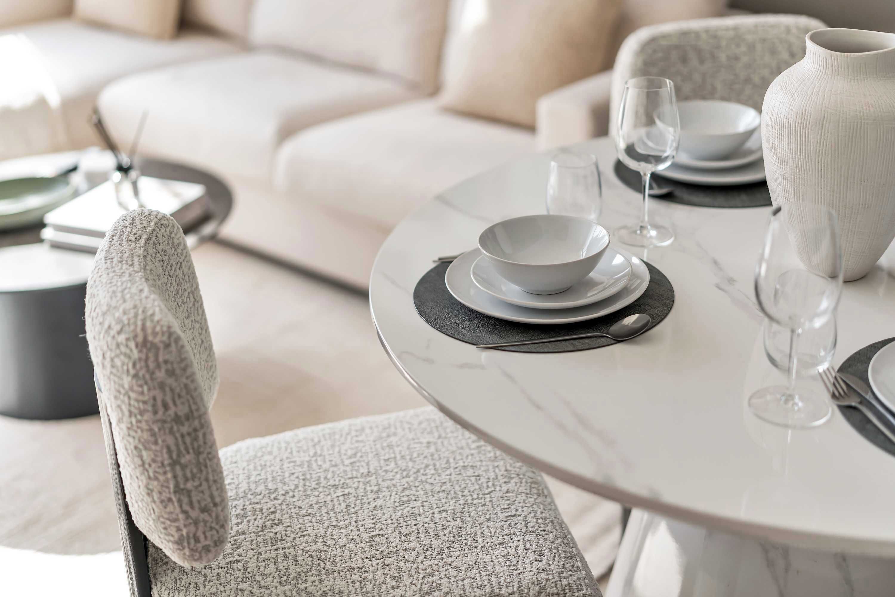 Uma mesa de jantar com pratos, tigelas e um vaso, disposta numa sala de estar moderna com um sofá e uma poltrona.