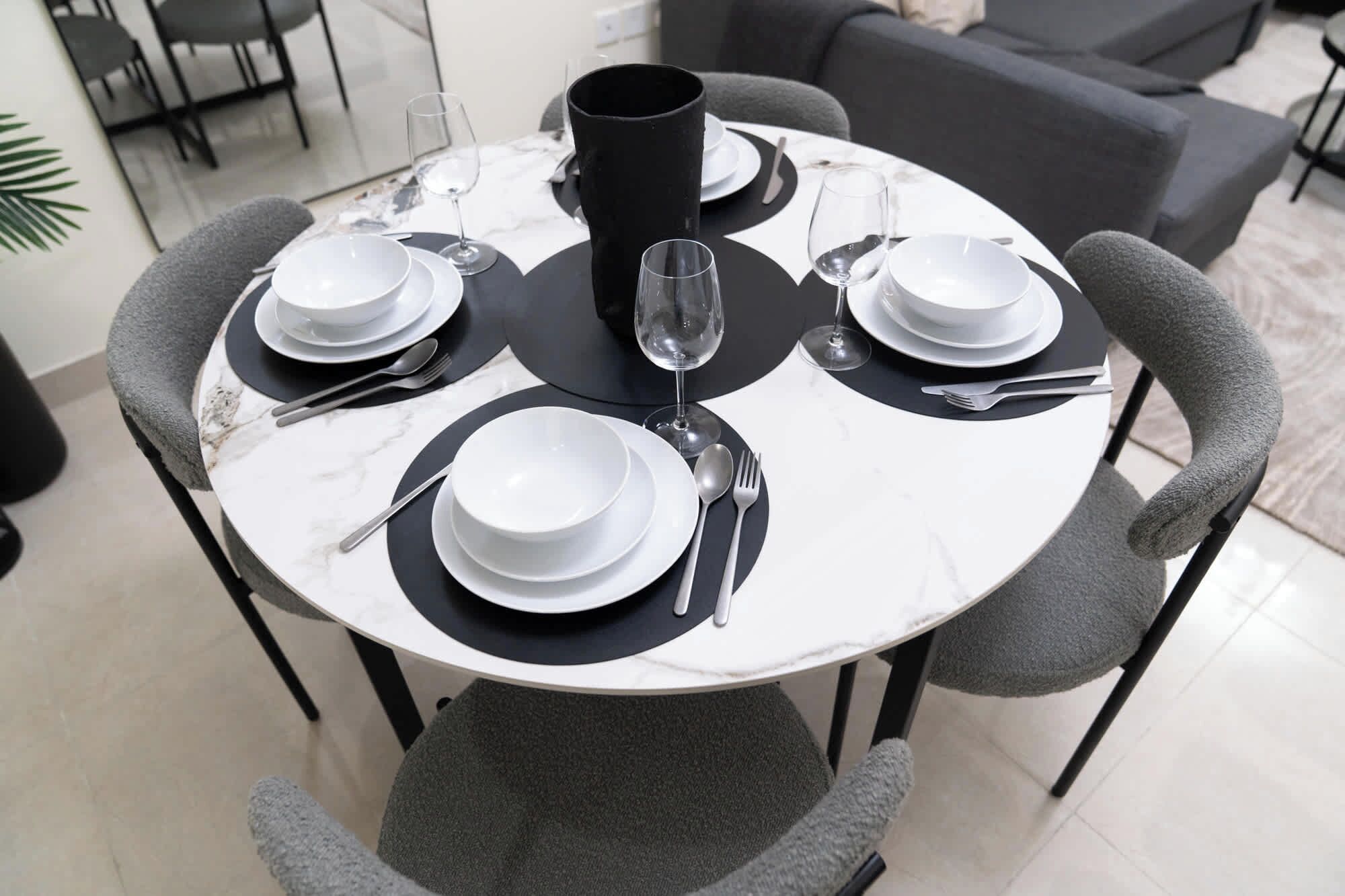 A round dining table with four chairs, each with a black cushion. The table is set with white plates, bowls, and silverware.