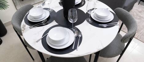 A round dining table with four chairs, each with a black cushion. The table is set with white plates, bowls, and silverware.