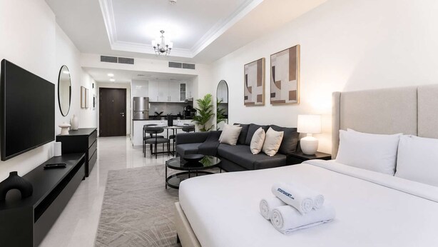 A modern hotel room with a large bed, a flat-screen TV, a sofa, a dining area, and a kitchen in the background. - Spacious Studio Apartment that Sleeps 4 (JVC)
