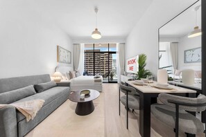 A modern living room with a sofa, dining table, and chairs, a large window with a city view, and a sign that reads 'BOOK ME'. - Free housekeeping visits during your stay - StayShort - Modern Studio that Sleeps 4 at Riviera Beachfront (Meydan)