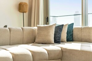 Ein modernes Wohnzimmer mit einem Ecksofa, einer Stehlampe und Blick auf eine Stadtkulisse durch große Fenster.