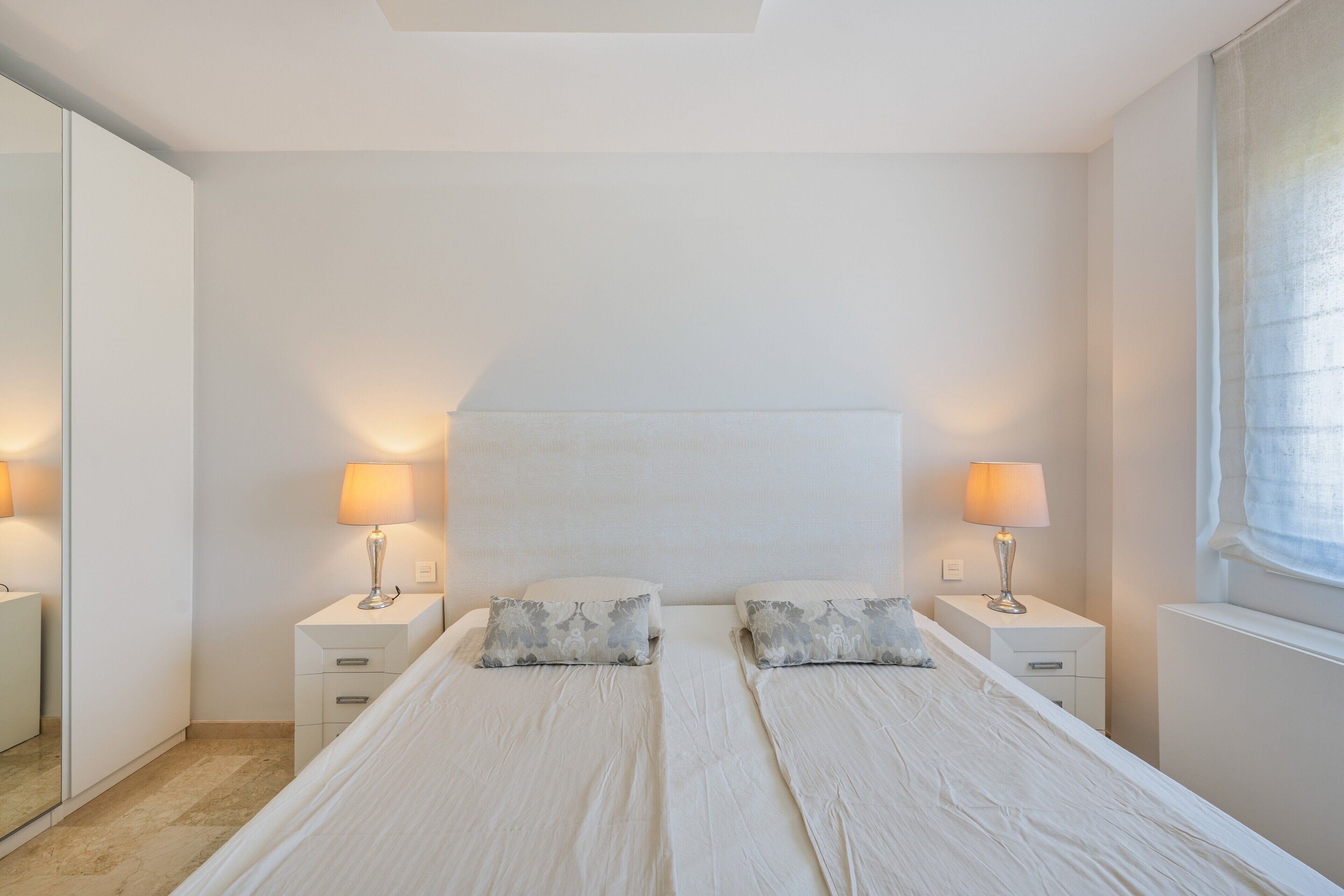 Une chambre à coucher avec un grand lit, deux tables de chevet avec des lampes, un miroir et une fenêtre avec des rideaux.