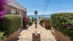 A narrow brick pathway leading to a building with a garage door, surrounded by lush greenery and a view of the sea.