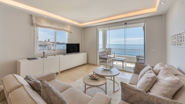 A modern living room with a sofa, a TV, a dining table, and a balcony with a view of the sea.