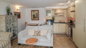 A living room with a sofa, a wooden table, and a kitchen with cabinets and a microwave. - Longfords Mill, Minchinhampton (Stroud)