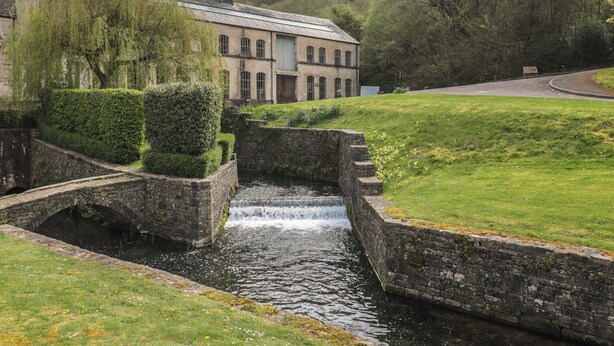 A historic building with a water feature, stone bridge, and lush greenery. - Longfords Mill, Minchinhampton (Stroud)