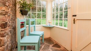 A room with two chairs, a small table, a hanging plant, and a view of greenery through large windows. - Whitefield Cottage (Barnstaple)